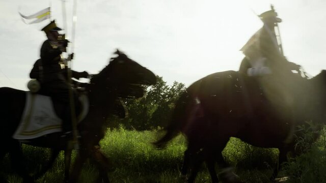 Polish cavalrymen wearing military uniform and holding lances riding horses in the field. Silhouettes of men and horses in slow motion.