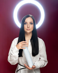 Dark-haired girl on a red background with a halo from a ring lamp and a bottle of hairspray in her hands