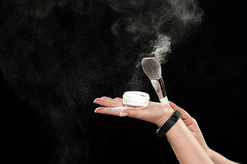 Female hands with powder and brush, powder beautifully dissolves through the air. Black background, close up, copy space. 