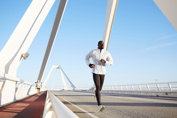 Full length portrait of a fit and athletic male running on a bridge road promenade outside - copy space area, dark skinned runner jogging against bright blue sky background.