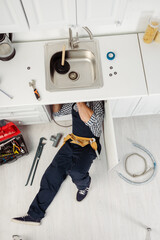 Top view of plumber in workwear fixing kitchen sink near tools on floor