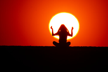 Young girl doing yoga exercises at sunset. In the background the big sun can be seen behind the silhouette of the girl. The concept of nature and beauty. Orange sunset.
