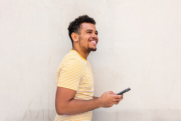 Side of happy young african american mixed race man by white wall holding mobile phone and looking away