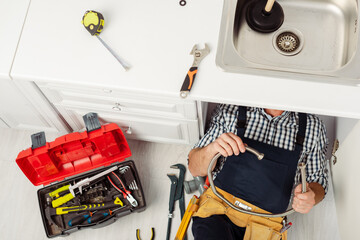 Top view of plumber holding metal pipe while repairing kitchen sink near tools on worktop and floor