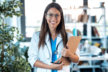 Happy young female doctor smiling and looking at camera while holding a clipboard and standing in...