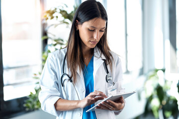 Confident young female doctor reviewing the patient's medical history on her digital tablet while standing in the consultation.