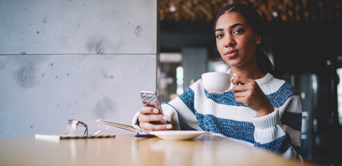 Attractive african female blogger enjoying tea time and communication with followers on content website via cellphone, portrait of beautiful woman looking at camera and drinking coffee in cafeteria