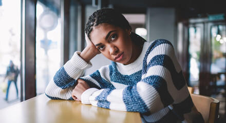 Portrait of attractive dark skinned hipster girl dressed in casual wear posing at table in university cafeteria, charming african woman looking at camera while waiting friend in cafe interior