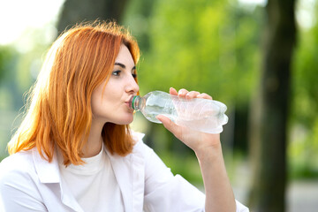 Young thirsty redhead woman drinking water from a bottle in summer park.