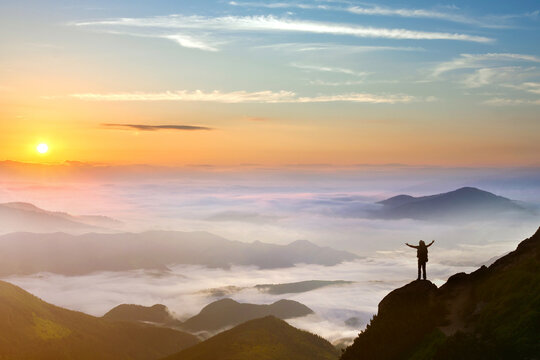 Small silhouette of hiker man enjoying beautiful sunrise in morning mountains. Traveler with raised hands standing on mountain with white fog below.