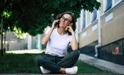 A phone call in the park. A full-length photo of a young girl, dressed casually, wearing the glasses on her face, sitting in the park, smilling, having her phone call.