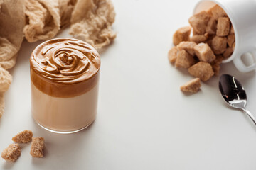 selective focus of delicious Dalgona coffee in glass near spoon, cup, cloth and granulated brown sugar on white background