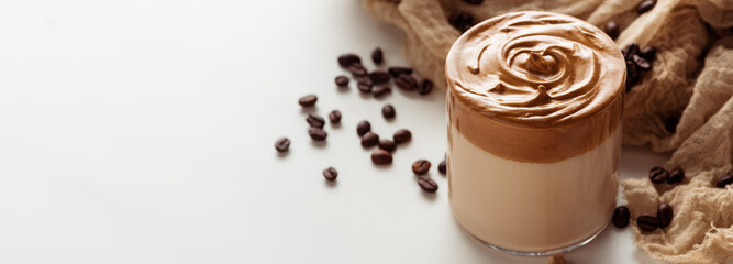 delicious Dalgona coffee in glass near coffee beans on cloth on white background, panoramic shot