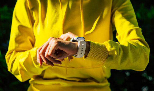 Modern device. A photo of a girl in a yellow hoodie wih yellow nails, with a white modern smartwatch on her hand, clicking on the touch screen.