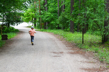 Little boy running away through green forest in summer day, back view. Children leisure outdoors activities.
