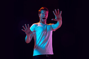 Palm greeting, scared. Caucasian young man's portrait on black studio background in neon light. Beautiful male model in white shirt. Concept of human emotions, facial expression, sales, ad. Copyspace.