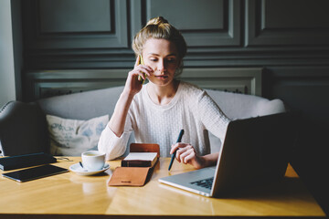 Thoughtful businesswoman having mobile conversation on telephone connected to wifi while looking at laptop computer.Charming blonde female working with modern devices sitting in cozy cafe