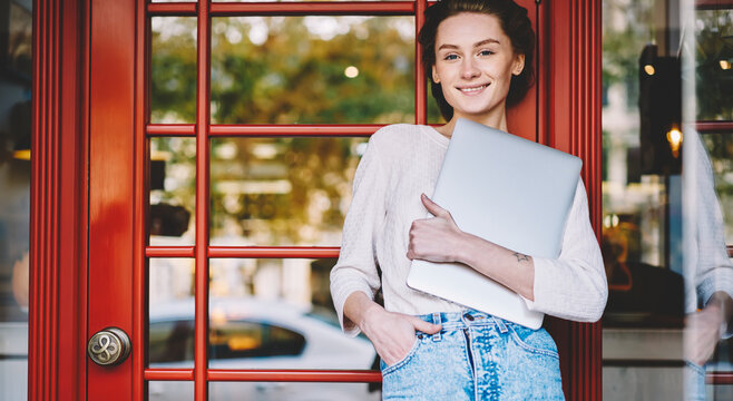 Portrait of positive female dressed in stylish casual clothing holding modern macbook computer in hand while looking at camera standing on promotional background.Smiling student with laptop device