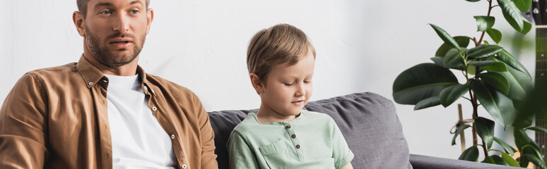 panoramic crop of thoughtful father and son sitting on sofa near potted plant