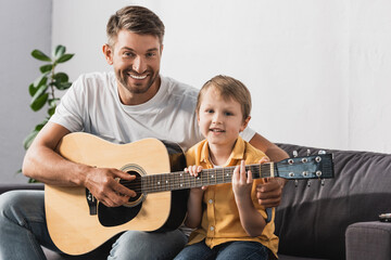 happy father teaching cheerful son how to play acoustic guitar