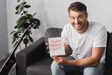 happy man showing happy fathers day card while sitting on sofa near plant and acoustic guitar