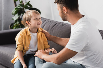 father touching shoulder of smiling son while talking to him on sofa