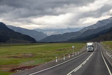 Fototapeta premium lorry on a highway in the mountains, stormy sky evening landscape, asphalt 2-lane road