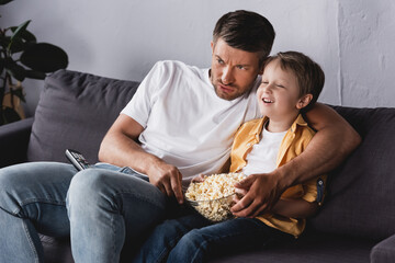 Obraz na płótnie Canvas worried father and smiling son watching tv and eating popcorn on sofa at home