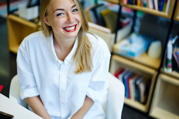 Cheerful attractive female student feeling happy completing successful coursework sitting in library near copy space area for advertising,carefree hipster girl in white shirt smiling sitting in office