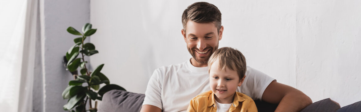 horizontal image of smiling father and son sitting on sofa near potted plant