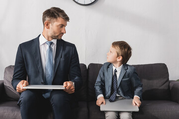 serious businessman and his son in formal wear sitting with laptops on sofa at home
