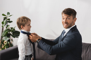 smiling businessman in formal wear looking at camera while putting tie on adorable son at home