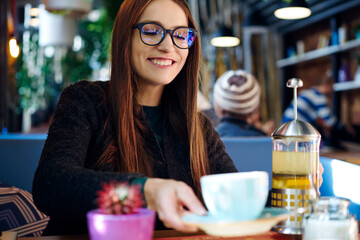 Cheerful foxy hipster girl in stylish eyeglasses sitting in cozy coffee shop with cup of taste tea and enjoying recreation time.Smiling young woman with red hair and trendy spectacles with good lenses
