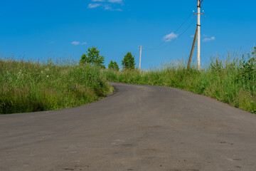 Fototapeta na wymiar An asphalt road stretching into the distance among grass, electric poles against a blue sky with small clouds.