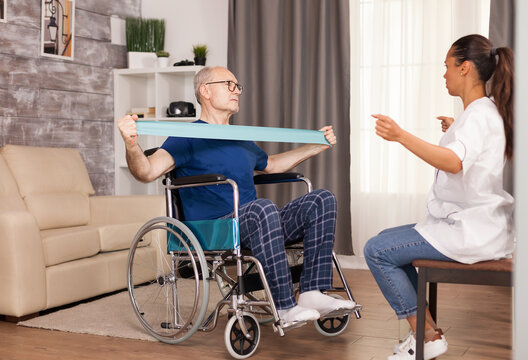 Invalid senior man in wheelchair training for muscle injury with therapist using resistance band. Disabled handicapped old person with social worker in recovery support therapy physiotherapy