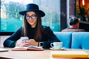 Smiling stylish hipster girl in eyeglasses and trendy hat reading funny sms message from friend on...
