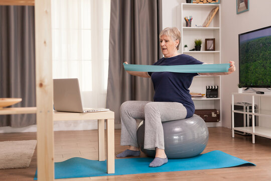 Elderly Woman Workout At Home In Front Of The Laptop. Old Person Pensioner Online Internet Exercise Training At Home Sport Activity With Dumbbell, Resistance Band, Swiss Ball At Elderly Retirement Age