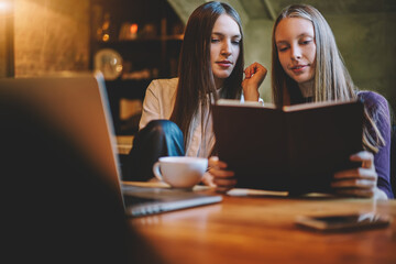 Intelligent female students reading training book and preparing together for upcoming exams sitting in stylish coworking space at modern laptop computer device connecting to wireless 4G internet