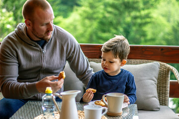 Father giving little son croissant for breakfast in the morning.Dad and his cute boy are talking and smiling while having breakfast on the terrace.Young happy father and son having breakfast together