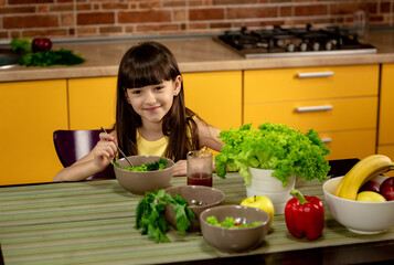 Little girl sitting at a table in the kitchen and eating a salad. Pretty little girl smiling. Healthy food, vegetables and fruits