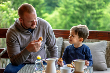 Father giving little son croissant for breakfast in the morning.Dad and his cute boy are talking and smiling while having breakfast on the terrace.Young happy father and son having breakfast together