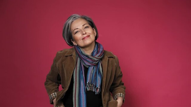 Gray-haired mature woman fixing her hair doing hairstyle. Creative Caucasian woman wearing casual clothes posing in studio on pink background. Prores 422. 