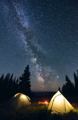 Tourist camp in the mountains, warm summer evening. Two illuminated tents and campfire burning under breathtaking night sky full of stars and shiny milky way. Concept of camping.