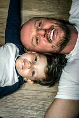 Top view of happy father and son close to each other heads and smiling while lying on the wooden floor. Close-up portrait of young dad and his son having time together. Family bonding.