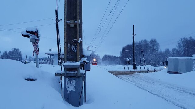 The road was blocked, a red warning light is on. Suburban train arrives. In the evening, the street lights were lit. Outdoors. Winter.