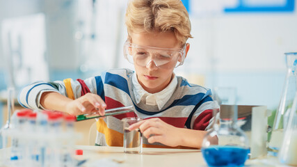 Elementary School Science or Chemistry Classroom: Smart Little Boy wearing Safety Glasses Mixes Chemicals in Beakers