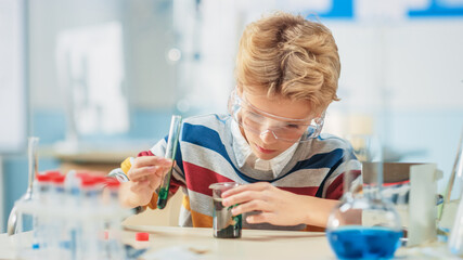 Elementary School Science or Chemistry Classroom: Smart Little Boy wearing Safety Glasses Mixes Chemicals in Beakers
