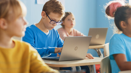Elementary School Computer Science Class: Smart Boy Uses Laptop Computer, His Classmates work with Laptops, Everybody Listen Attentively to the Teacher. Children Getting Modern Education