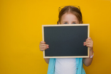 Fototapeta na wymiar Portrait of a beautiful little girl peeking from behind a blackboard in her hands, smiling on a yellow background. Attractive positive baby. back to school. Copyspace