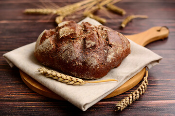 Dark rye bread on a linen napkin on a light wooden board and ears of wheat on a brown wooden table. Rustic still life.